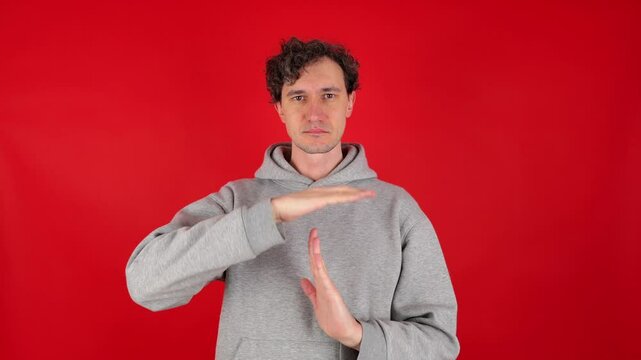 young adult curly serious man holds hands in front of shows sign timeout gesture. wearing grey hoodie. isolated red background studio shot. Concept - pause, break, rest, vacation