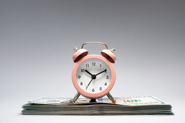 A pink alarm clock sits atop a tidy stack of cash, illustrating the connection between time...