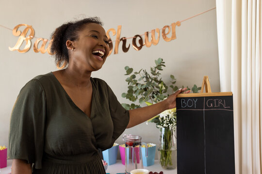 African American woman laughing at baby shower with gender reveal board, at home
