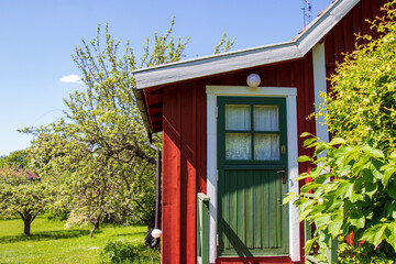 A classic red wooden cottage in Stockholm, Sweden with a green door, surrounded by greenery and fruit trees under a bright blue sky.