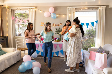 Diverse female friends celebrating baby shower with decorations and gifts in living room, at home