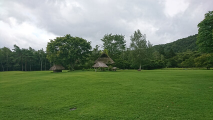 Naklejka premium 曇り空の中、山や木々を背景に広大な緑の芝生の公園の中央に茅葺き屋根の小屋が点在する風景