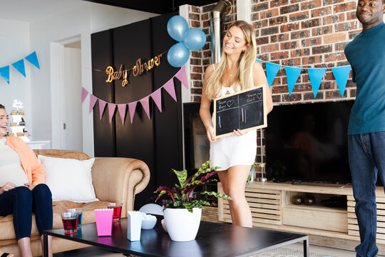 Joyful woman holding chalkboard at Baby shower celebration with diverse friends at home
