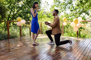 Man proposing to surprised woman on wooden deck surrounded by balloons