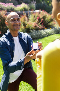 Man proposing with ring box in garden, smiling and kneeling on grass