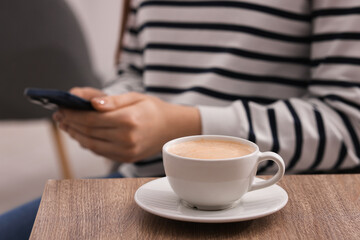 Woman using smartphone during coffee break at wooden table indoors, closeup