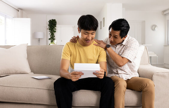 Asian father congratulating son reading acceptance letter on couch at home