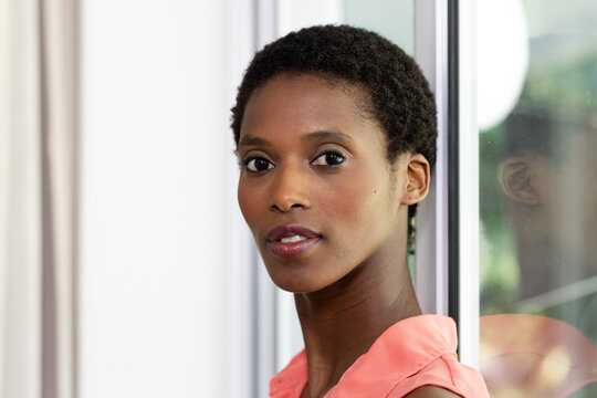 Confident woman in coral blouse standing by window, looking thoughtfully ahead, at home, copy space