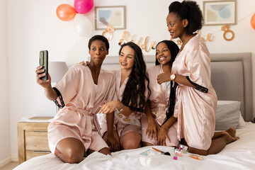 Bachelorette party, diverse female friends in matching robes taking selfie on bed during celebration