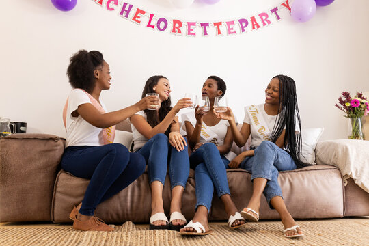 Celebrating bachelorette party, diverse female friends toasting with drinks on sofa, at home