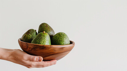 A person is holding a bowl of avocados. The avocados are green and the bowl is wooden