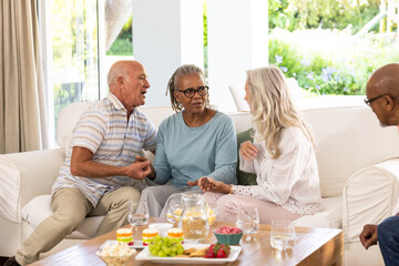 Senior diverse friends enjoying conversation and snacks together in cozy living room, at home