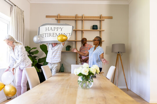 Senior diverse friends decorating room with balloons and banner for retirement celebration, at home