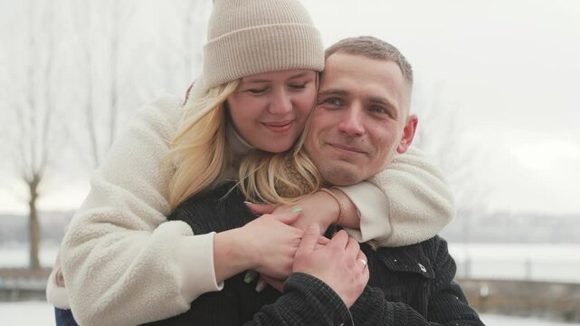 A happy couple in love is having a great time in a nearby winter park. The woman is hugging and tenderly kissing the man.