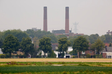 Obraz premium Industrial landscape featuring smokestacks near residential area with surrounding greenery on a hazy day