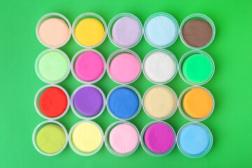 Different colorful modeling clay in plastic jars on green background, flat lay