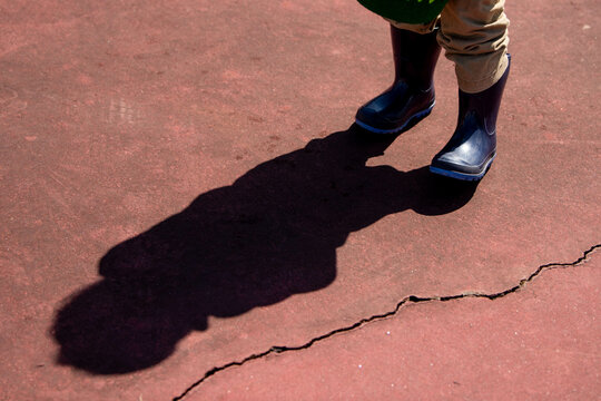 A child's shadow stretches out across cracked red pavement. The child is wearing blue rubber boots and wrinkled brown pants. 