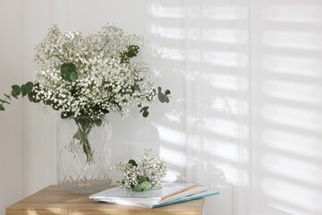 Beautiful gypsophila flowers with eucalyptus branches in vase and magazines on table at home. Space...