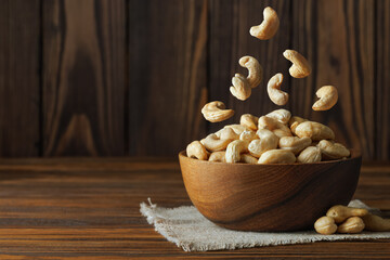Cashew nuts falling into bowl placed on small rustic jute tablecloth, creating a visually appealing arrangement against dark wooden background