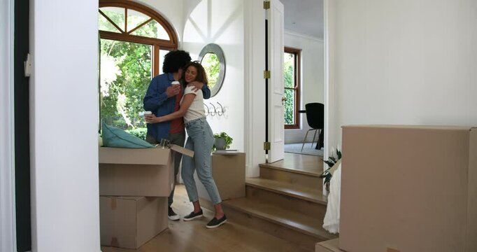 Excited diverse couple with coffees, walking between boxes, hugging and looking around new home