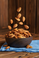 Shelled raw almonds falling into bowl placed on blue napkin on rustic wooden table, creating warm...