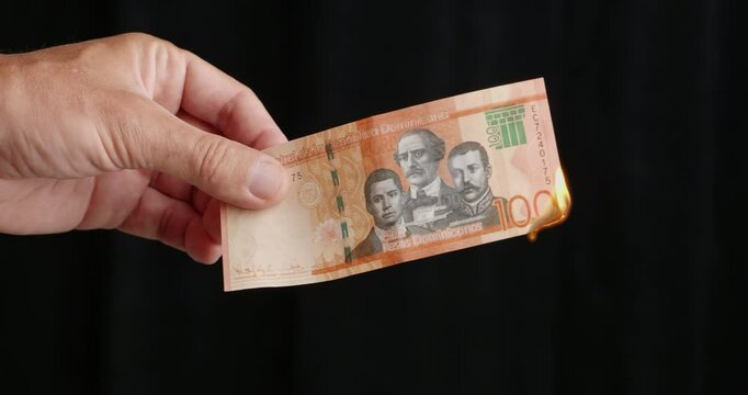 Burning hundred Dominican peso banknote. Hand holds the banknote while lights it on fire using a lighter. Inflation concept. 