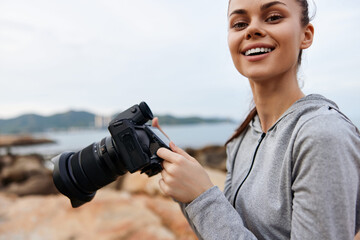 Young woman holding a camera by the seaside, showcasing joy and confidence in a casual outfit,...