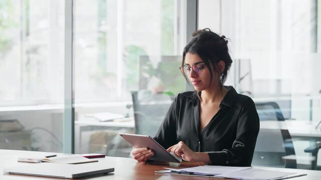 Latin Hispanic young professional business woman working on touchscreen pc computer. Middle eastern Indian businesswoman CEO holding digital tablet, using tab pad application sitting at desk in office