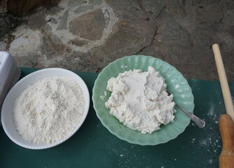 Ingredients and equipment ready to make cretan traditional xortopitakia - Two bowls on a table: one with white flour, the other with mizythra, a cretan cheese, in a light green, scalloped dish 