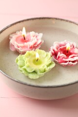 Beautiful burning flower shaped candles in dish with water on pink wooden table, closeup