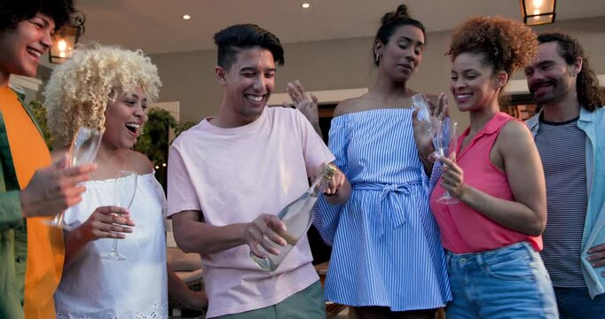 Happy diverse friends opening champagne, talking and pouring drinks to celebrate at a party