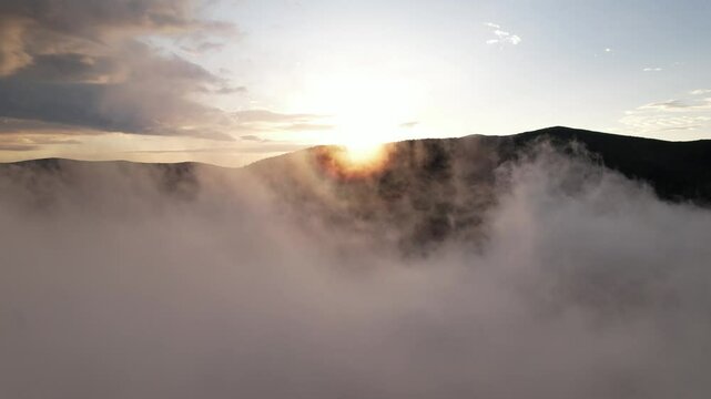Aerial shot of a sun rising from the ridge of a mountain with a flying through the cloud