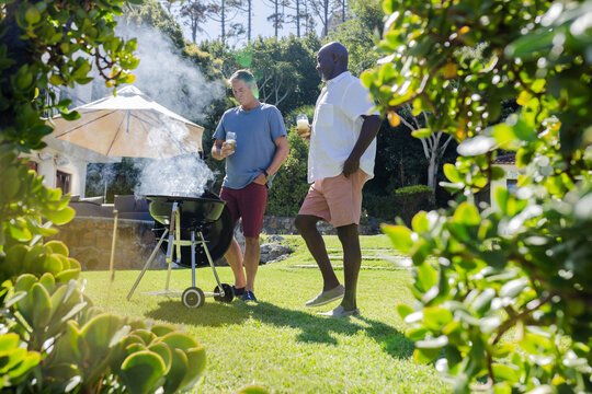 Senior friends enjoying outdoor barbecue, holding drinks and chatting in sunny backyard