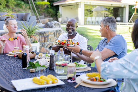 In garden, diverse senior friends sharing barbecue food, laughing together outdoors