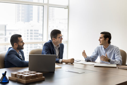 Three professionals engaged in discussion seated at conference table in modern office, listening male leader explaining or presenting new idea. Formal business meeting event or brainstorming session
