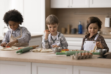 Friendly cooking. Busy team of three multicultural boys friends preschoolers engaged in preparing meal at home kitchen cook homemade bakery cookies preparing pastry from dough together for snack lunch