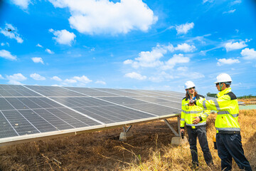 Two engineers wearing safety helmets and reflective jackets inspect a solar panel installation. One uses a drill tool, while the other checks the panel surface under a bright, sunny sky.