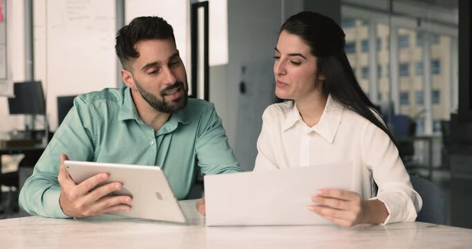 Couple of young Latin project colleagues analyzing reports on tablet and in paper documents, discussing financial information, comparing data, consulting each other, giving professional assistance
