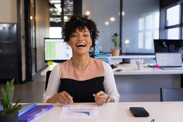 African American woman laughing at office desk with documents and smartphone