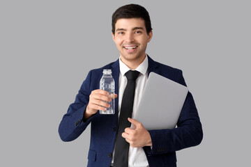Young businessman with bottle of water and laptop on grey background