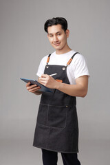 Confident Asian man wearing an apron and using a digital tablet and stylus, demonstrating focus and...