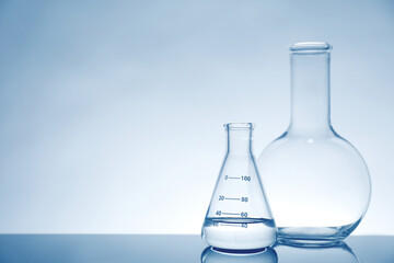 Laboratory glassware with liquid on mirror surface against light background, space for text