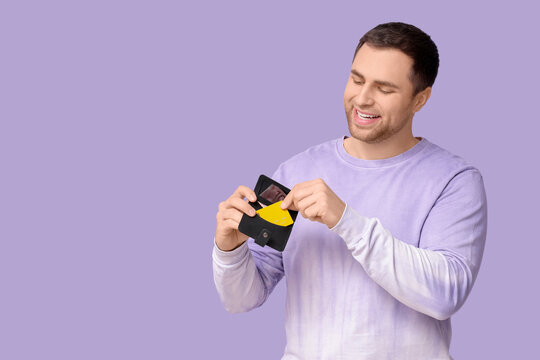 Young happy man putting credit card in wallet on lilac background