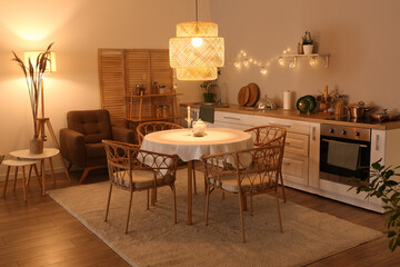 Interior of modern kitchen with glowing lamps, dining table, oven and white counters at evening