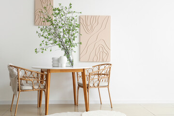 Table, armchairs and vase with blossoming branches near white wall in beautiful dining room