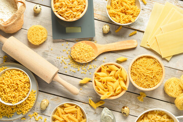 Different types of raw pasta and kitchen utensils on light wooden background