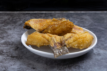 Crispy fried fish served on a white plate against a dark background, showcasing its golden-brown coating and flaky texture