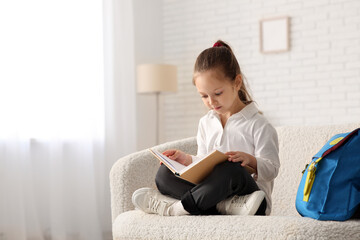 Cute little girl with book and backpack sitting on sofa and studying at home