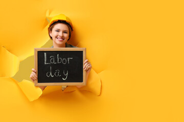 Young female carpenter holding chalkboard with text LABOR DAY through hole in yellow paper. Labor...
