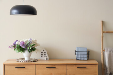 Vase with lilacs on wooden chest of drawers and lamp in stylish room, closeup